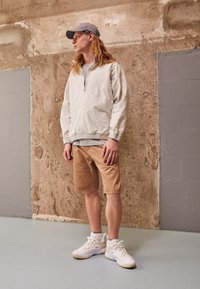 Lys beige pullover med en kvart lynlås, tan shorts og hvide sneakers. Afslappet design og løs pasform, sat mod en tekstureret væg.