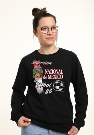 Kvinde med briller iført en sort sweatshirt med teksten "Seleccion Nacional de Mexico futbol 86", fodboldemblem og grafik af en bold.