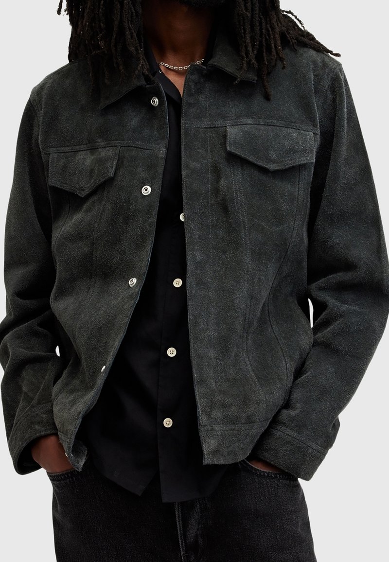 Mörkgrön mockajacka med knäppning fram, två bröstfickor och lätt struktur. Bärs över en svart skjorta och kombineras med mörka jeans.