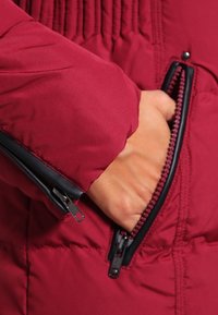 Sezione di una giacca imbottita bordeaux con tasca con zip visibile, dotata di accessori neri e di un tessuto testurizzato, evidenziando una mano inserita nella tasca.