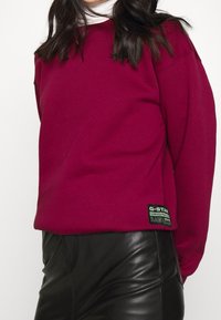 Burgunderfärgad sweatshirt med rund halsringning, sänkta axlar och en grön etikett med varumärkesinformation. Matchas med svarta läderbyxor.