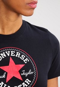 Svart bomullströja med en röd stjärnlogotyp där texten "Converse" och "Chuck Taylor" är i vitt, placerad mot en rund bakgrund.