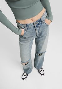 Light blue denim jeans with distressed knee rips, straight leg design, five-pocket style, and silver-tone button closure. Black and white sneakers.