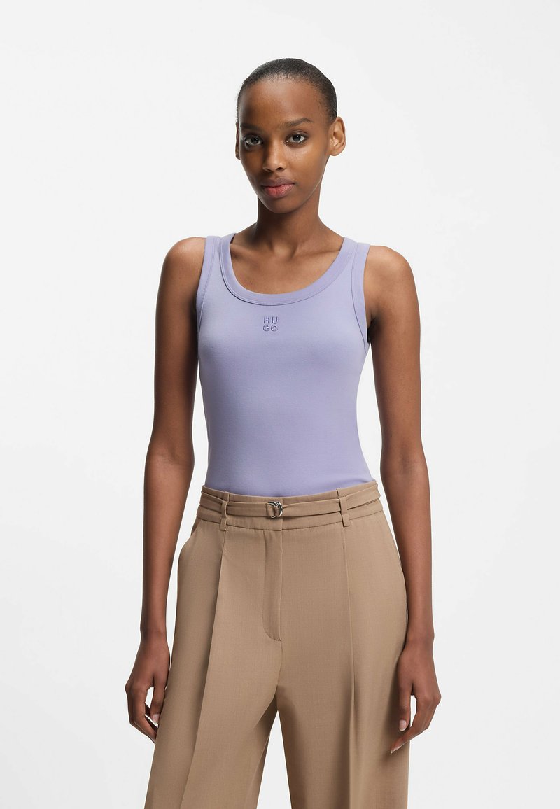 Lilaaninsävyinen, vartalolle istuva toppi pyöreällä pääntiellä, jossa on hillitty "HUGO"-logo. Yhdistetty korkeavyötäröisiin beigen värisiin housuihin, joissa on vyöhön liittyvä yksityiskohta.