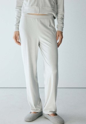 Persona que lleva pantalones de descanso gris claro, parte superior con cremallera a juego y zapatillas grises y peludas, de pie frente a un fondo liso.