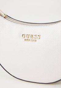Borsa bianca strutturata con forma curva, hardware dorato e chiusura a cerniera. Presenta il logo in rilievo "GUESS EST 1981" sul davanti.