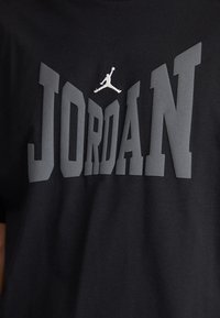 Juodas medvilnės marškinėliai su dideliu pilku "JORDAN" užrašu ir balta Jumpman logotipu virš jo. Švelni tekstūra su šiuolaikiniu bloko stiliaus šriftu.