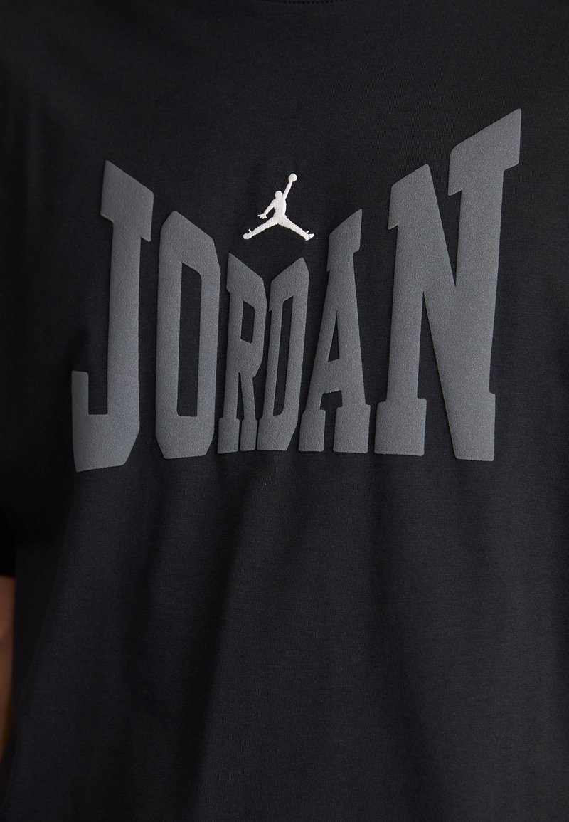 Czarna bawełniana koszulka z dużym szarym napisem "JORDAN" oraz białym logo Jumpman powyżej. Miękka tekstura z nowoczesnym blokowym krojem pisma.