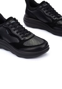 Zapatillas deportivas negras que presentan una combinación de cuero suave y acentos perforados, con una suela gruesa y elementos de diseño texturizados.