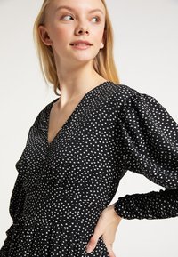 Rochie neagră cu mici modele de inimioare albe, decolteu în V, mâneci bufante și un corset ajustat care se transformă într-o fustă evazată. Textură moale a materialului.