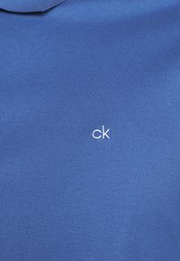 Tissu de coton bleu à surface texturée, avec un petit logo blanc "ck" sur le côté gauche.