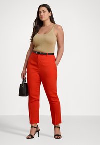 Canottiera a coste marrone chiaro, pantaloni rossi a vita alta, cintura nera e tacchi, che tiene una piccola borsa nera. Sfondo neutro.
