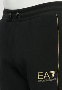 Un pantalon de survêtement noir présente une finition texturée, une taille élastique avec cordon de serrage, et un logo doré EA7 Emporio Armani sur la cuisse gauche.