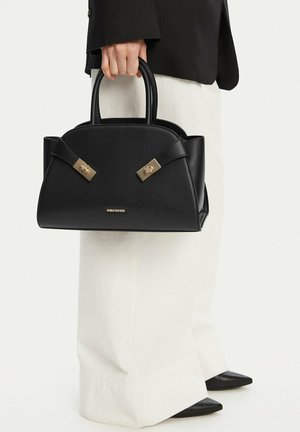 Persona che tiene una borsa nera Gino Rossi con dettagli dorati, indossando pantaloni bianchi a gamba larga, scarpe nere appuntite e una giacca nera.