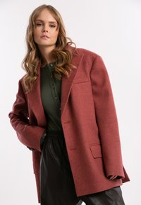 Przesadny, wełniany blazer w odcieniu różowo-czerwonym, z klapami w stylu notched, jedną kieszenią na piersi oraz guzikami przy mankietach. Nosi się go na zielonym, prążkowanym topie.