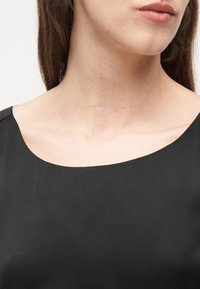 Κοντινό πλάνο στο κάτω μέρος του προσώπου και στον λαιμό μιας γυναίκας που φοράει μαύρο τοπ, με μια αχνή, οριζόντια ουλή να φαίνεται στον λαιμό της πάνω από το κόκαλο του αυχένα.