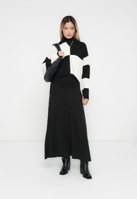 Pulôver de malha preto e branco cortado com meia abertura, combinado com uma saia longa preta e botas pretas de salto grosso, e uma mala preta.