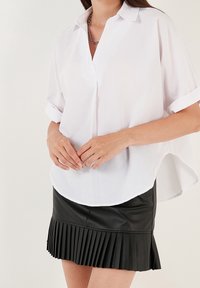 Weißes kurzärmeliges Hemd mit Kragen, weicher Textur und abgerundetem Saum, kombiniert mit einem schwarzen, plissierten Minirock aus Kunstleder.