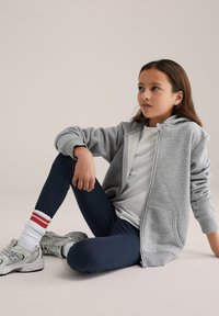 Szürke cipzáras kapucnis pulóver, fehér póló, tengerészkék leggingsek, piros csíkos zoknik és szürke sportcipők. Puha anyag, laza szabás és első zsebes kialakítás.