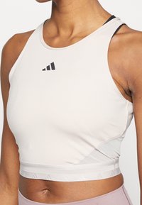 Světle růžový sportovní crop top s přiléhavým střihem, který má černé logo a strukturovaný lem. Vyrobeno z hladkého, elastického materiálu.