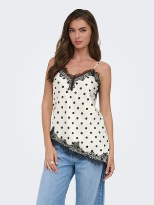 Kvinde iført en hvid satin camisole med sorte polkaprikker og blonder, kombineret med lyseblå jeans med vide ben, stående foran en hvid baggrund.