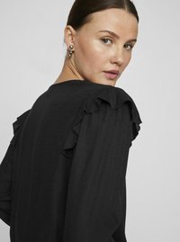 Crna bluza s volanima na ramenima, teksturiranim materijalom i opuštenim krojem. Dizajn ima okrugli izrez i duge rukave.