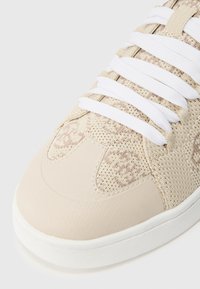 Μπεζ αθλητικό παπούτσι με λευκά κορδόνια, με πλεκτό ύφασμα και συνθετικά επικαλύμματα, εμφανιζόμενο σε κοντινή λήψη στην περιοχή των δακτύλων.