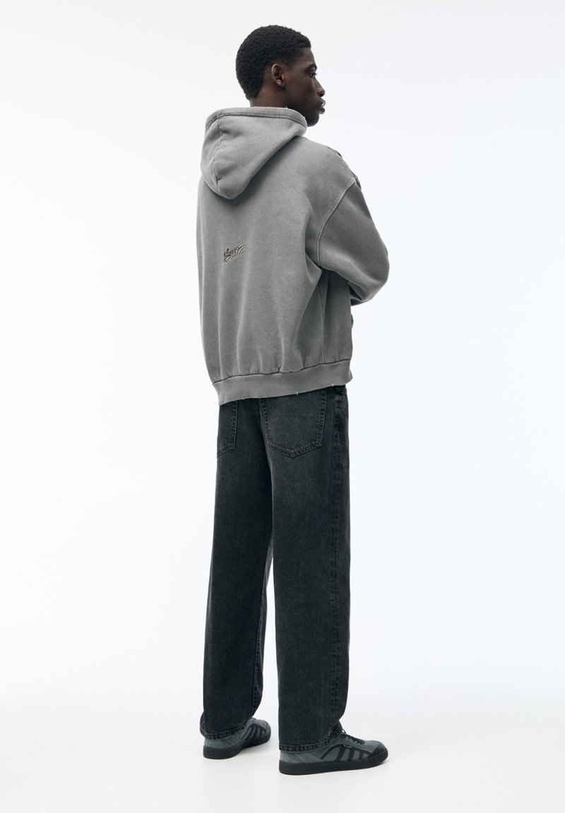 Felpa grigia con cappuccio e orlo elastico, abbinata a jeans a gamba larga grigi scuri. Calzature nere e un design minimalista in evidenza.