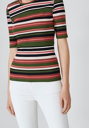 Kobieta ubrana w dopasowany, prążkowany top z krótkim rękawem w poziome paski w kolorach zielonym, koralowym, czarnym i białym, w zestawieniu z białymi spodniami.