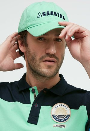Man adjusting light green Gaastra cap, wearing matching navy and green polo with Gaastra Sailing Heritage logo patch.