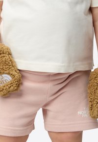 Beež värvi lühikeste varrukatega T-särk otse lõigatud äärega, mida on kombineeritud helepunaste lühikeste pükstega. Laps hoiab pehmeid pruune karvaseid kinnastikuid, millel on valge logo.