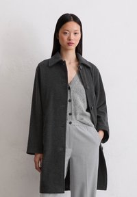 Vrouw in een donkergrijze button-up jas over een lichtgrijze cardigan en bijpassende lichtgrijze broek, staand tegen een effen gestructureerde muur.