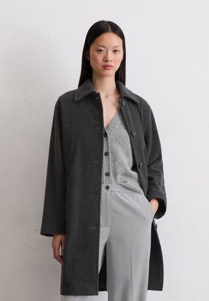 Femme portant un manteau boutonné gris foncé par-dessus un cardigan gris clair et un pantalon gris clair assorti, se tenant devant un mur texturé uni.