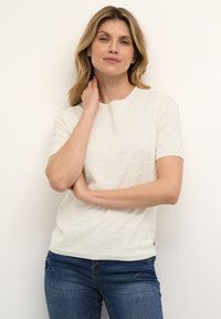Creme kortærmet sweater med teksturerede bobble-detaljer, rund halsudskæring og tætsiddende pasform, parret med blå jeans.