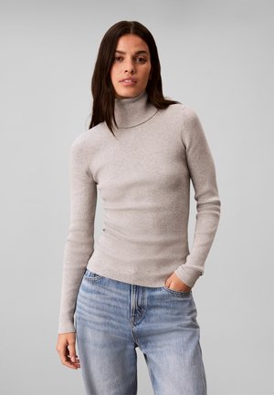Femme aux longs cheveux foncés portant un pull à col roulé côtelé gris clair et un jean bleu clair, une main dans la poche.