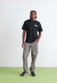 Black cotton t-shirt with a small pink logo, paired with grey relaxed-fit pants and black sneakers, standing on a green floor.