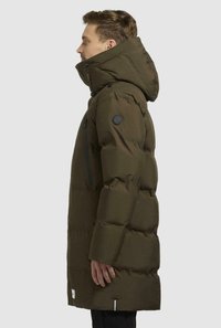 Cappotto imbottito verde oliva con colletto alto, cappuccio rimovibile e patch con logo. Presenta una tasca sul petto con zip e tasche laterali.