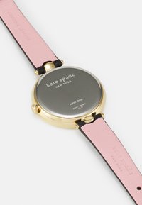 Guldtonad klocka med rund urtavla, svart och rosa läderrem, och ingraverat varumärke på baksidan. Slät metallisk yta.