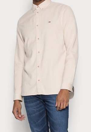 Mand iført en lys beige skjorte med knapper og en lille logo på brystet og blå jeans, der står mod en ensfarvet baggrund.