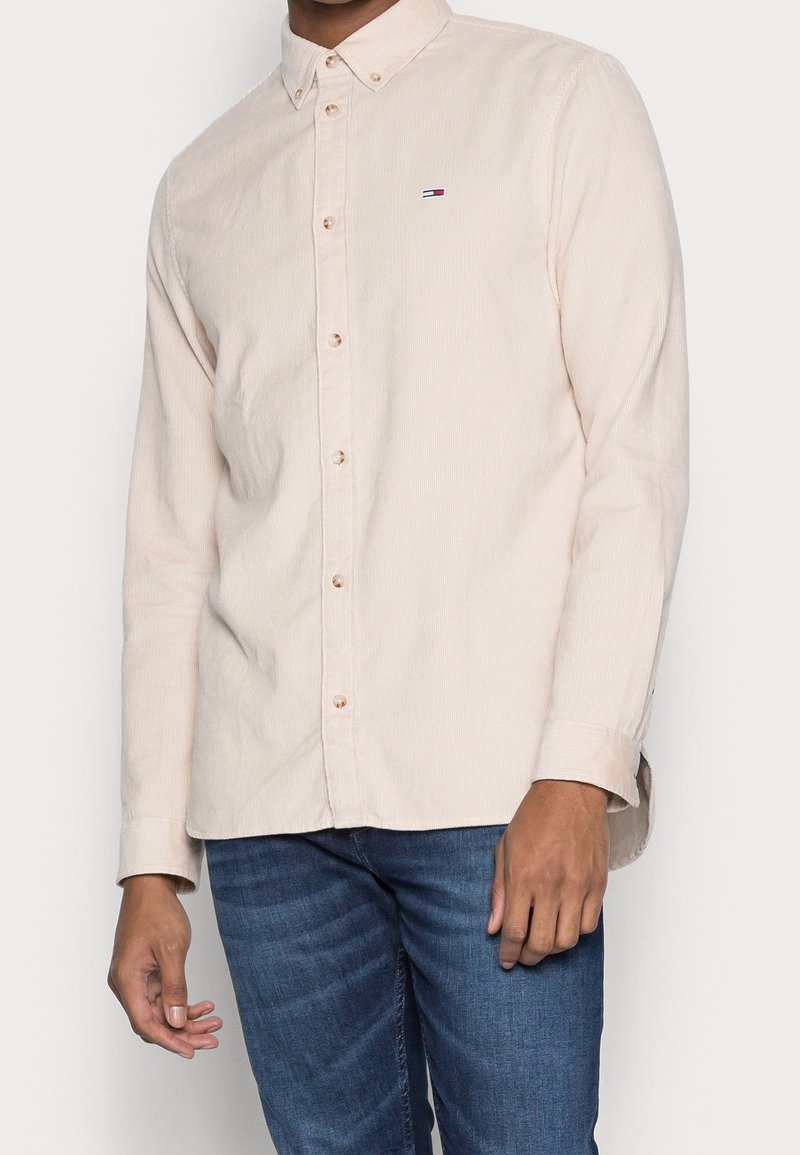 Homme portant une chemise beige clair à boutons avec un petit logo sur la poitrine et un jean bleu, debout devant un fond uni.