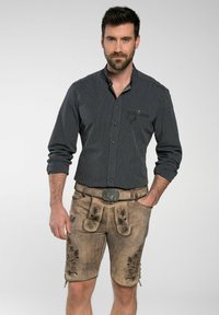 Ein Mann trägt ein schwarz-weiß gestreiftes Hemd mit Knöpfen und beige Leder-Shorts mit floralen Stickereien sowie einer dekorativen Gürtelschnalle.