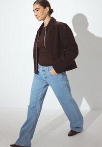 Giacca in suede marrone scuro con vestibilità cropped, zip in argento e colletto, abbinata a jeans a campana blu chiaro e scarpe marrone scuro.