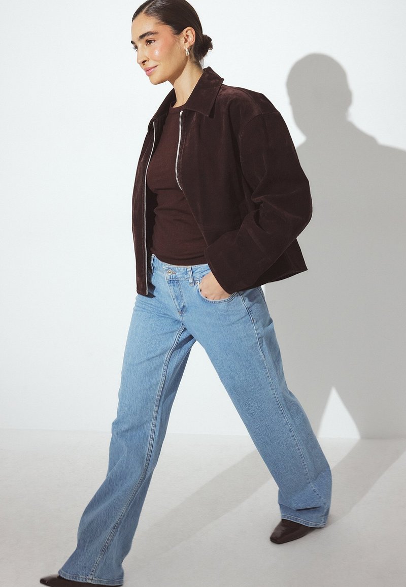 Giacca in suede marrone scuro con vestibilità cropped, zip in argento e colletto, abbinata a jeans a campana blu chiaro e scarpe marrone scuro.