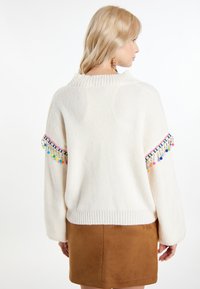 Suéter de punto crema con ribete de pompones de colores en las mangas, cuello y dobladillo acanalados, combinado con una falda de ante marrón.