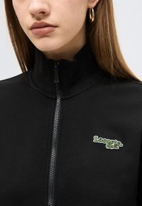 Νεαρή γυναίκα που φοράει μαύρο μπουφάν με φερμουάρ και το λογότυπο της Lacoste, καθώς και χρυσούς κρίκους σκουλαρίκια, εμφανιζόμενη από τον λαιμό έως το μέσο του στήθους.