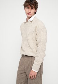 Bézs, kötött, zsinóros pulóver kerek nyakkivágással, texturált mintákkal. Fehér galléros ing fölött viselve, barna tweed nadrággal párosítva.