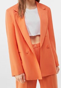 Oranžový dvouřadý blazer s klopy, vyrobený z hladké látky. Má dva knoflíky a boční kapsy, doplněný cropped bílou košilí.