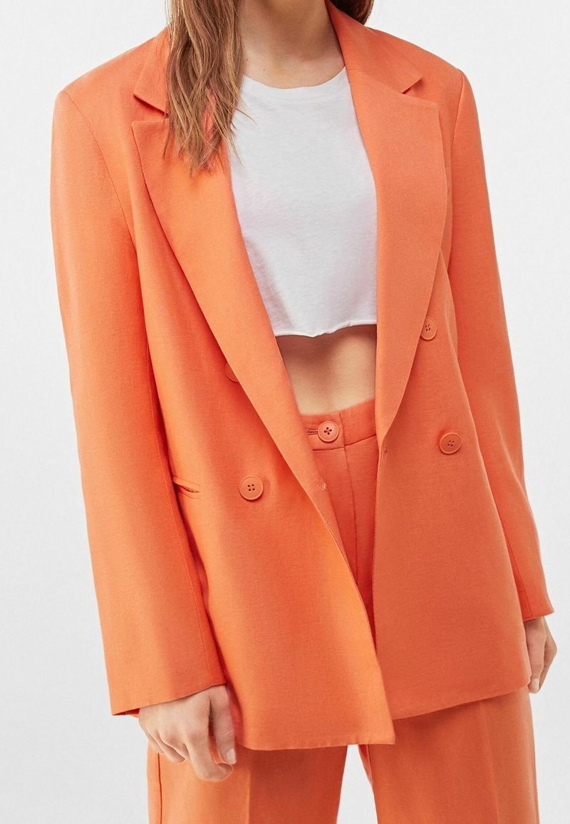 Oranžový dvouřadý blazer s klopy, vyrobený z hladké látky. Má dva knoflíky a boční kapsy, doplněný cropped bílou košilí.