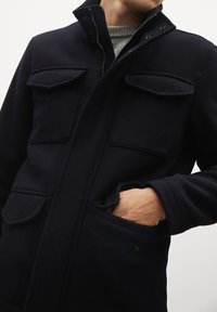 Mies, joka on pukeutunut tummansiniseen takkiin, jossa on neljä läpällistä taskua ja vetoketjukiinnitys. Hänen kätensä on osittain alhaisen oikean taskun sisällä, ja harmaa neule on näkyvissä.