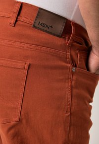 Närbild på rostfärgade byxor med synlig bakficka och läderetikett med texten "MEN+" nära midjebandet.